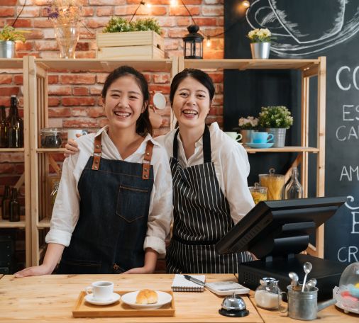 Two people in aprons behind a café counter with coffee, pastry, and notebook on the counter.